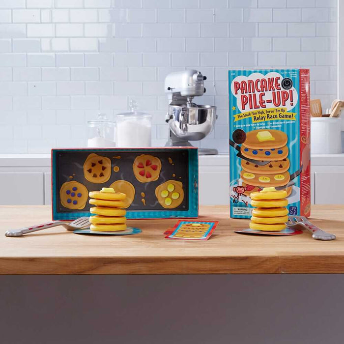 Yellow toy pancake stacks with butter, spatulas, and game box on a wooden counter against a white tile backsplash.