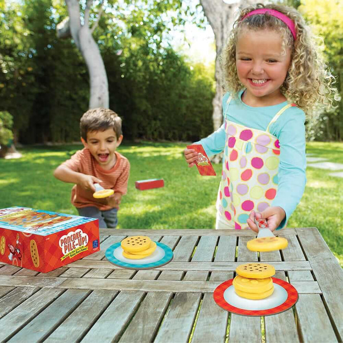 Children use spatulas to stack yellow toy pancakes on colorful plates outdoors next to a red game box on a wooden table.