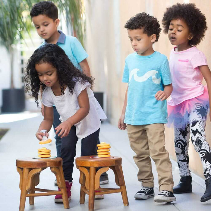 Four children play a relay game stacking yellow toy pancakes on wooden stools using small plastic spatulas.