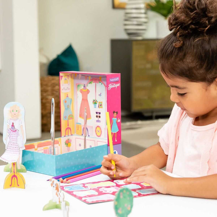 Girl colors a paper craft boutique set featuring a pink and blue shop box, paper dolls, and colored pencils.