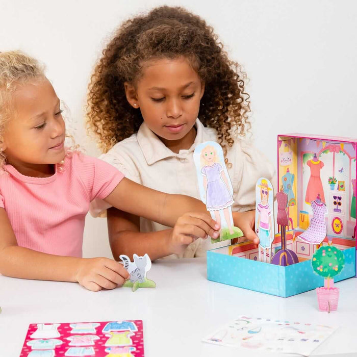 Two girls play with paper dolls and a pink fold-out shop with colorful cutout clothes and a cat on a white table.