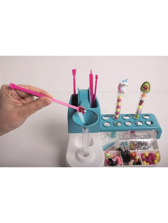 Hand fills a tube at a blue craft station with beads, charms, and custom unicorn and avocado topped pens.