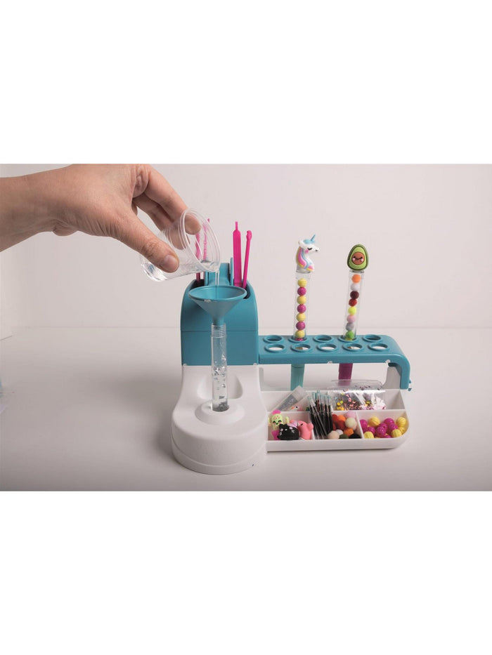 Hand pouring liquid into a funnel on a blue and white craft station with colorful beads and character pen toppers.