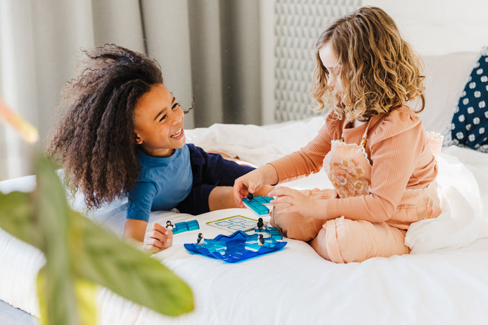 Two children play with sliding translucent blue ice blocks and small penguin figures on a white bed.