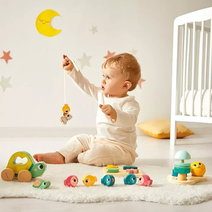 Toddler playing with wooden magnetic fishing rod, turtle pull toy, and colorful fish on a white rug.