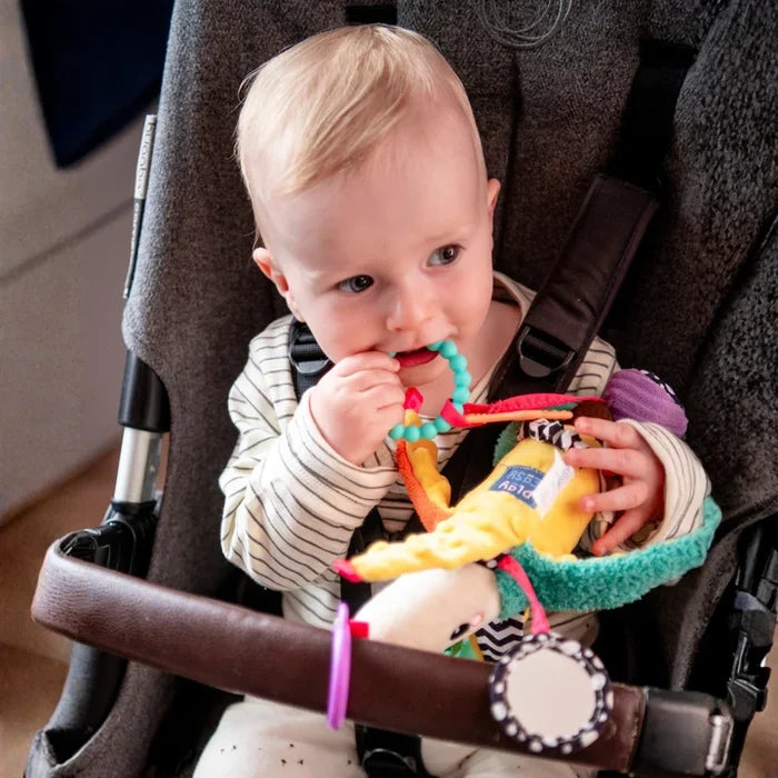 Baby in a stroller chewing on a yellow plush banana toy with colorful teether rings and textured sensory attachments.