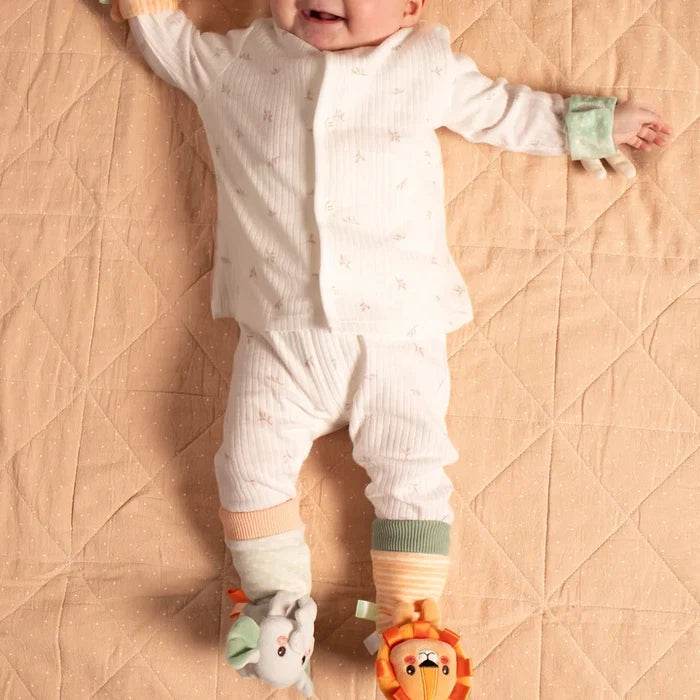 Baby in white pajamas on a quilted mat wearing plush lion and elephant foot rattles and a green wrist rattle.