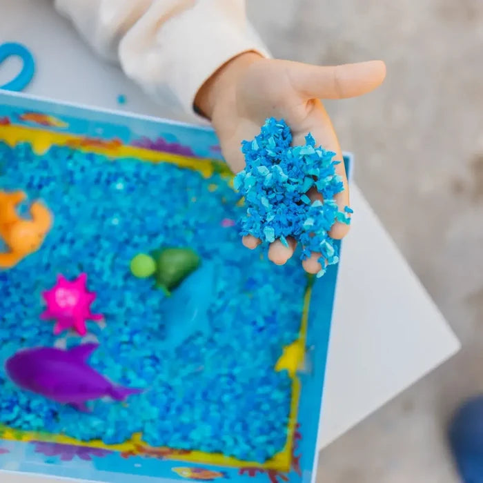 Hand holding blue fluffy foam over an ocean sensory bin with colorful sea creatures like a purple shark and pink starfish.