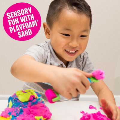Smiling boy playing with bright pink, blue, and green sensory sand using a roller and scoop toy on a white table.