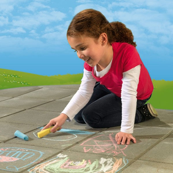 Young girl kneeling on a patio and drawing with yellow sidewalk chalk, with blue chalk on the ground nearby.