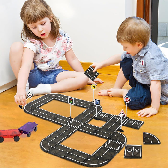 Two children play with a figure-eight track made of black magnetic road tiles and toy signs on a wooden floor.