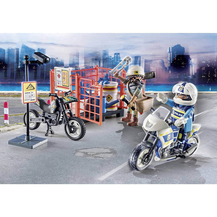 Toy police officer on white bike waves to a worker near a black dirt bike and orange storage cage with a barrel.