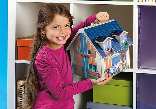 Girl in a magenta shirt holds a portable dollhouse with a blue roof and handle near white storage cubbies.
