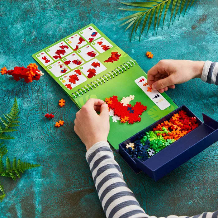 Child building a red dinosaur with plus-shaped blocks on a green activity pad next to a tray of colorful pieces.