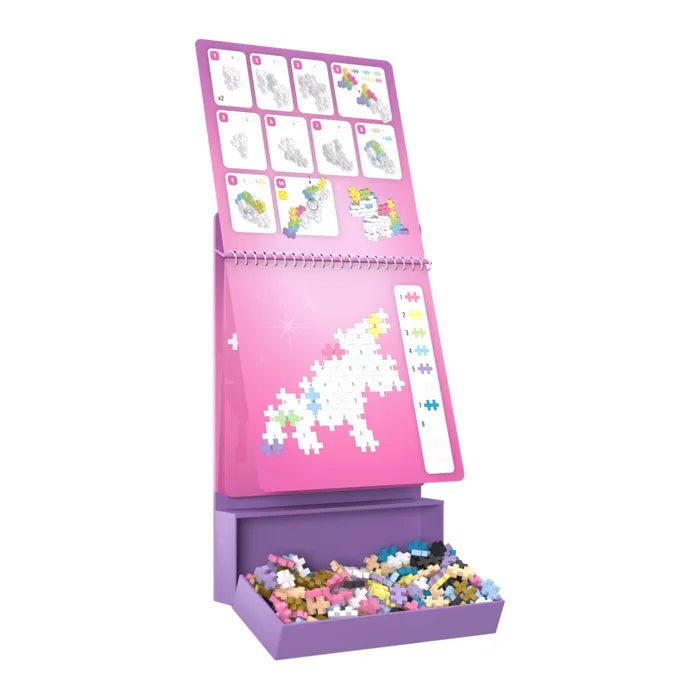 Pink spiral-bound unicorn activity pad on a purple easel with a tray of colorful cross-shaped building pieces.