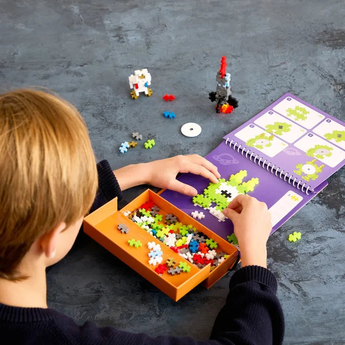 Child builds a green alien on a space activity pad with colorful plus-shaped blocks and an orange storage tray.