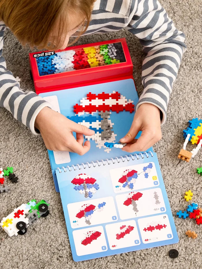 Child builds colorful transportation models using plus-shaped blocks on a spiral activity pad with a red storage tray.