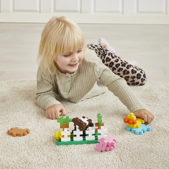 A blonde child playing on a rug with large colorful cross-shaped blocks arranged into a farm scene with animals.