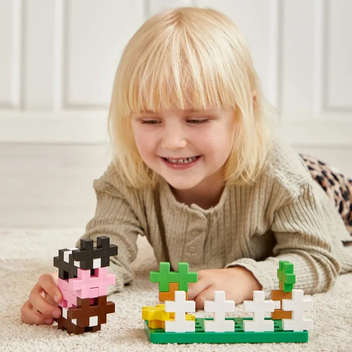 Smiling blonde girl builds a farm scene with large colorful plus-shaped blocks on a light carpet.