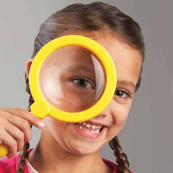 A smiling girl holds a large, bright yellow plastic magnifying glass over her eye, enlarging it.