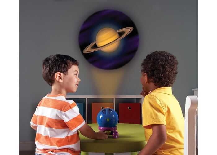 Two boys use a small blue globe toy to project a large, bright image of Saturn onto the wall behind them.
