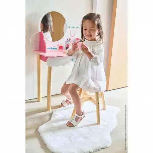 Young girl sitting at a pink and white wooden vanity with round mirror, stool, and play accessories on a white rug.