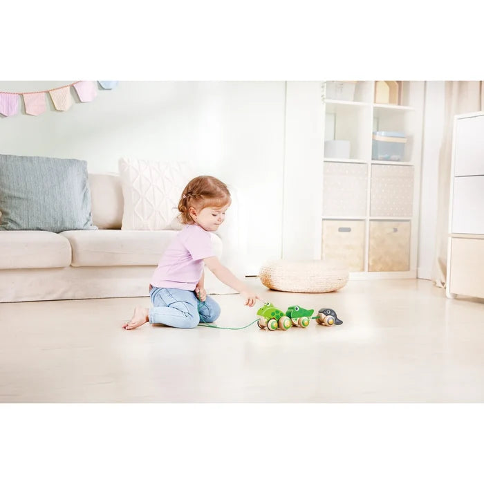 Toddler points at three wooden green and black pull-along frogs on wheels connected by a string on a light floor.