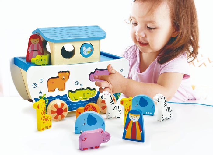 Toddler playing with a colorful wooden boat shape sorter and matching animal figurines on a white background.