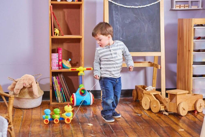 Toddler pulling three colorful blue, green, and yellow plastic ducks on wheels by a string across a wooden floor.
