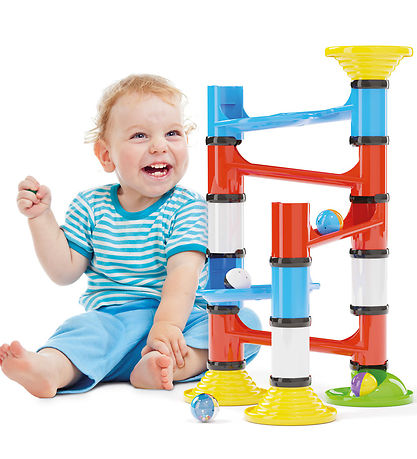 A toddler sits next to a colorful plastic ball track with chunky red, blue, and white tubes and large rolling balls.
