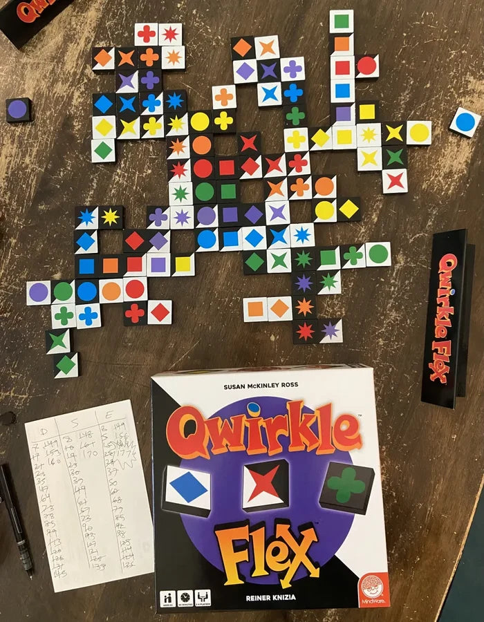 Qwirkle Flex game box with colorful symbol tiles arranged on a table next to a score sheet and tile rack.