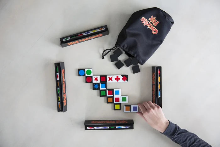 Overhead view of Qwirkle Flex being played with colorful shape tiles, black tile racks, and a branded drawstring bag.
