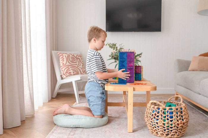 A child builds a tall tower with colorful, translucent magnetic rectangles on a wooden coffee table.