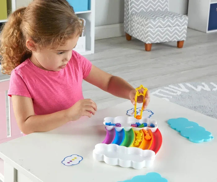 Child uses yellow star tongs to sort colorful pieces into a rainbow-shaped tray with white cloud compartments.
