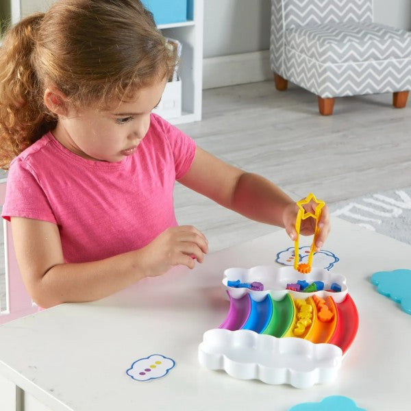 A child uses yellow star tongs to sort colorful shapes into a matching rainbow tray with white cloud ends.