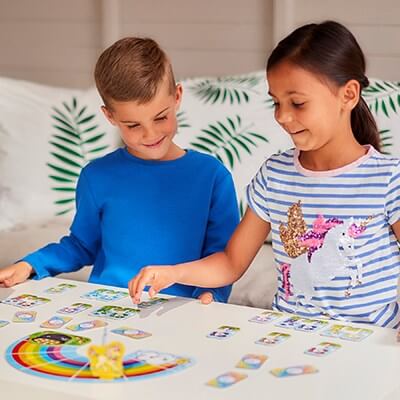 Two children smile while playing with colorful unicorn cards and a rainbow board piece on a white table.