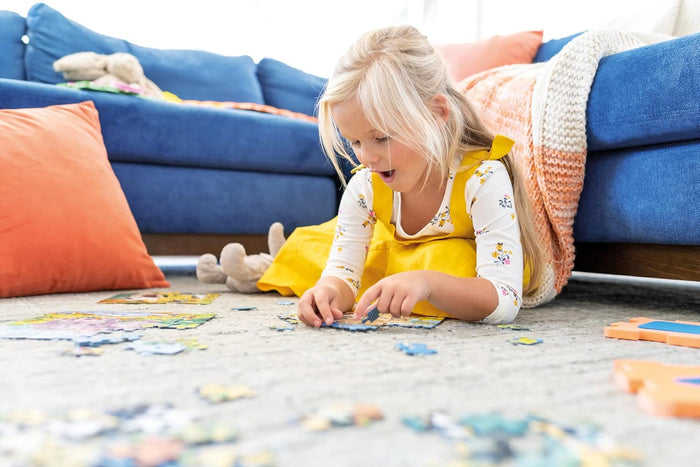 A young girl in a yellow dress excitedly assembles colorful jigsaw puzzle pieces on a gray rug near a blue sofa.