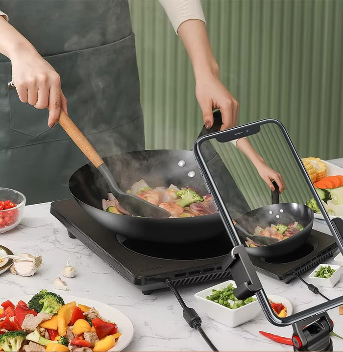 Person cooking vegetables in a wok while filming with a phone mounted on a tripod on the kitchen counter.