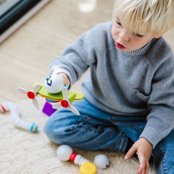 Blonde child building a green magnetic toy plane with white propellers and blue eyes on a textured rug.