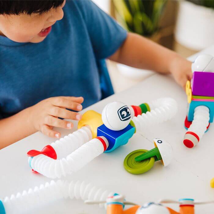 Child builds colorful magnetic robots with flexible white tubes, bright cubes, and round heads on a white table.