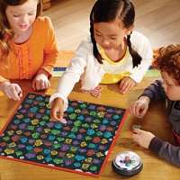 Three children playing a board game with a black board covered in colorful robot heads and a round shaker device.