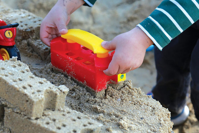 Child using a red plastic sand brick mold with a yellow handle to create textured bricks in a sandbox.