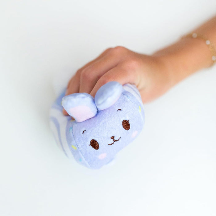 Hand squeezing a light purple bunny-themed Swiss roll plush with an embroidered face and colorful sprinkle details.