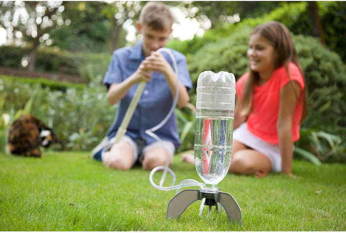 A boy pumps air into a clear plastic water rocket on a grassy lawn as a girl watches.