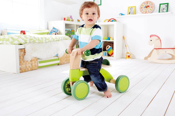 Toddler on a lime green and light wood four-wheeled ride-on toy with round green handlebars in a bright room.