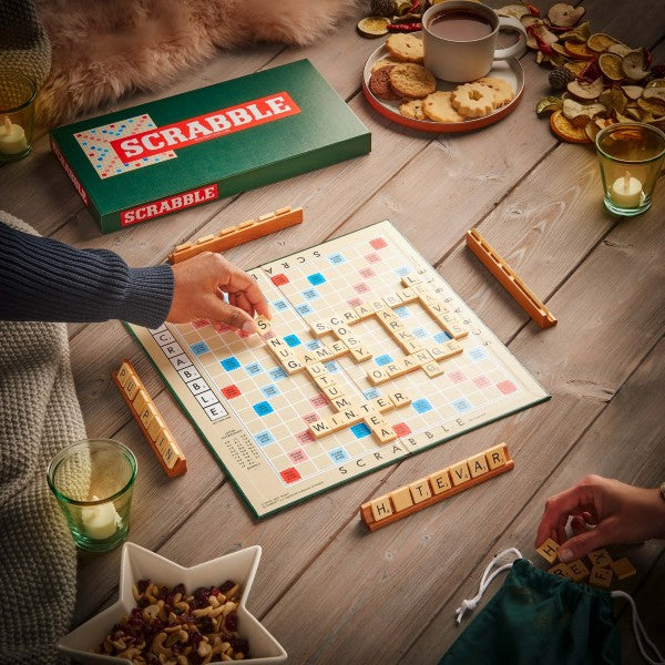 Scrabble board game on a wooden table with a green box, letter tiles, and cozy snacks like tea and cookies.