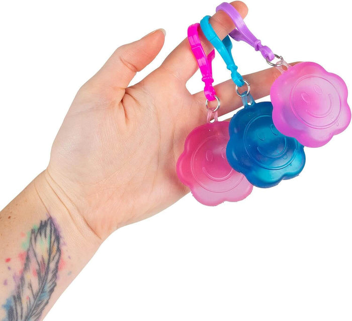 Hand holding three translucent, flower-shaped squishy keyrings with smiley faces in pink, blue, and purple.