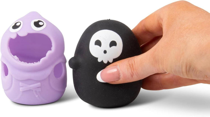 Hand squeezing a black skull-faced squishy toy next to a purple ghost shell with googly eyes and an open mouth.