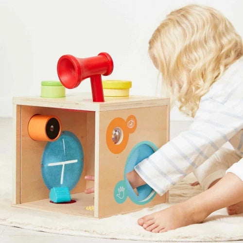 Child reaching into a wooden sensory cube toy with a red horn, blue oval, and colorful cutouts for touch and sight.