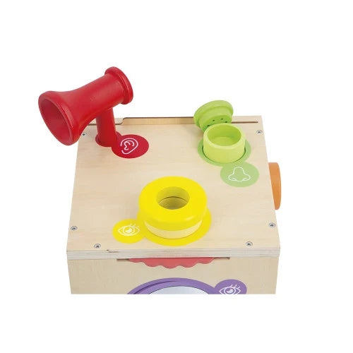 Wooden activity box featuring a red horn, green lidded jar, and yellow ring with icons for hearing, smell, and sight.
